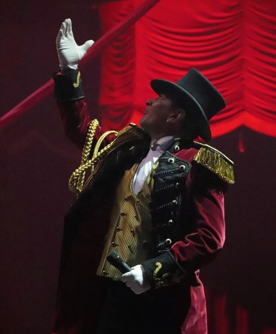 Homme dans un cirque, vêtu d'un gilet doré, une veste rouge à revers noirs et épaulettes or, coiffé d'un haut de forme et paré de gants blanc.