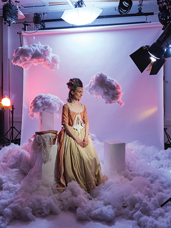 Femme prise en photo dans un studio, vêtue d'une robe couleurs or et bronze, dans un décor empli de décorations en forme de nuages.