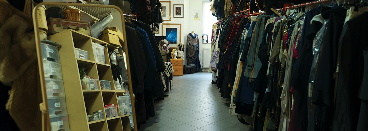 Allée des réserves de costumes, comprenant à droite des tenues sur un rack, au fond un mannequin habillé, et à gauche au premier plan, un miroir.