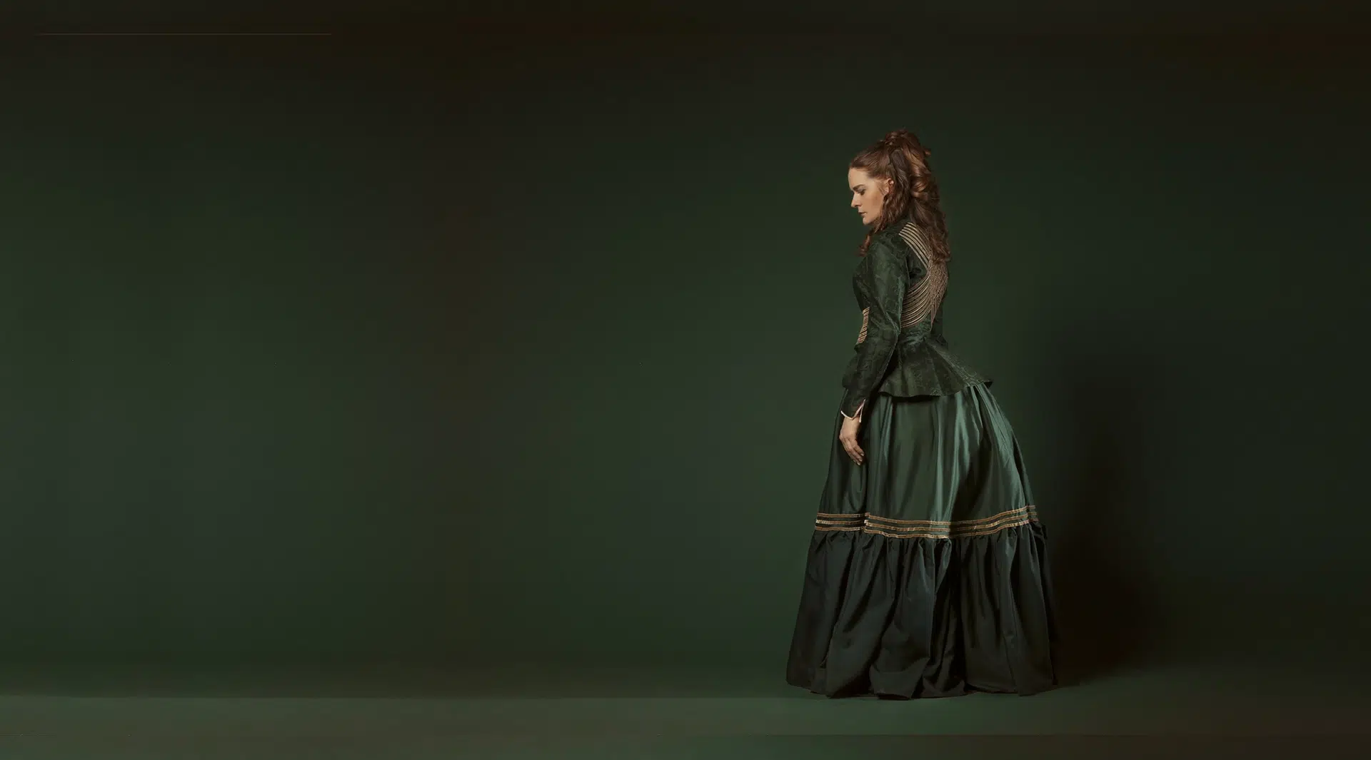 Femme de profil, vêtue d'une robe verte constituée d'une robe orné de trois bandes or et enflée d'une tournure, et d'un corset paré de bandes or au dos.